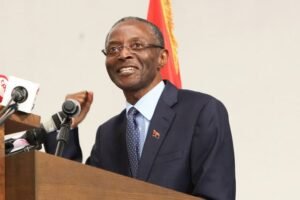 Homem de fato e óculos discursa diante de microfones e da bandeira de Angola.