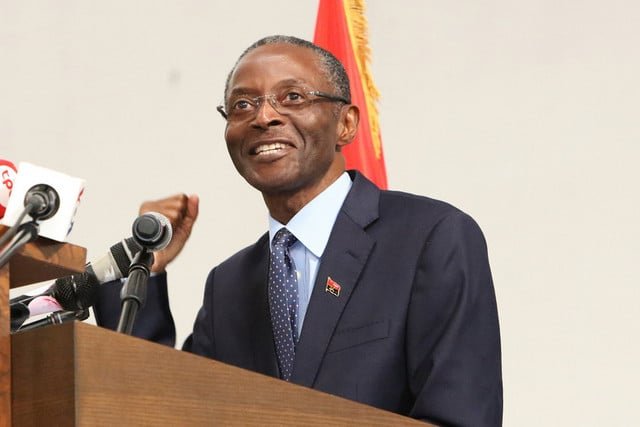 Homem de fato e óculos discursa diante de microfones e da bandeira de Angola.