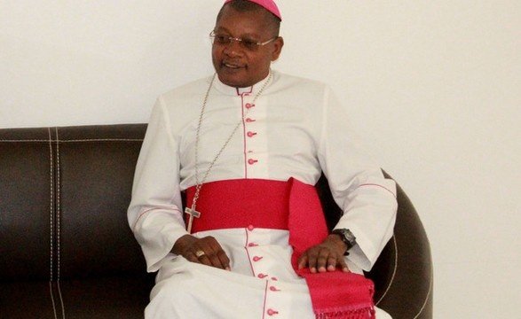Bispo sentado, com vestes brancas, faixa vermelha e solidéu.
