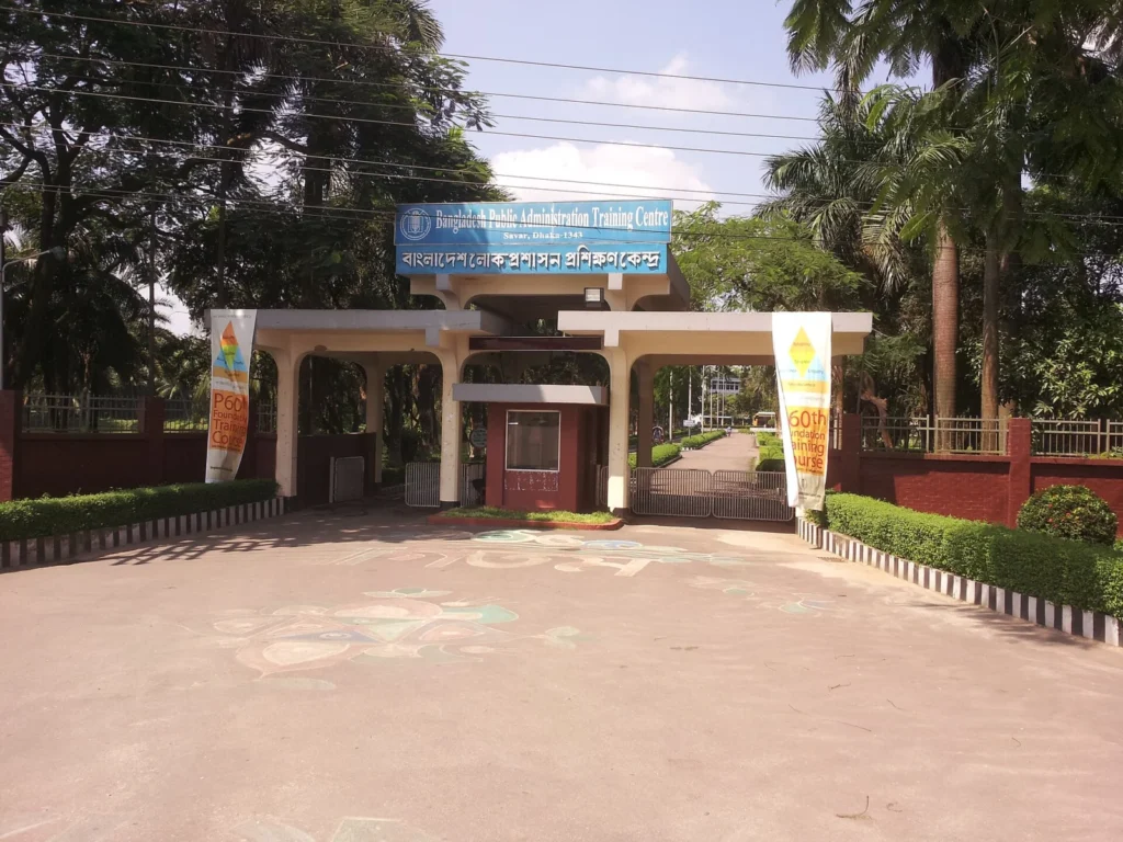Entrada do Centro de Formação em Administração Pública do Bangladesh, com portão, placa e árvores.