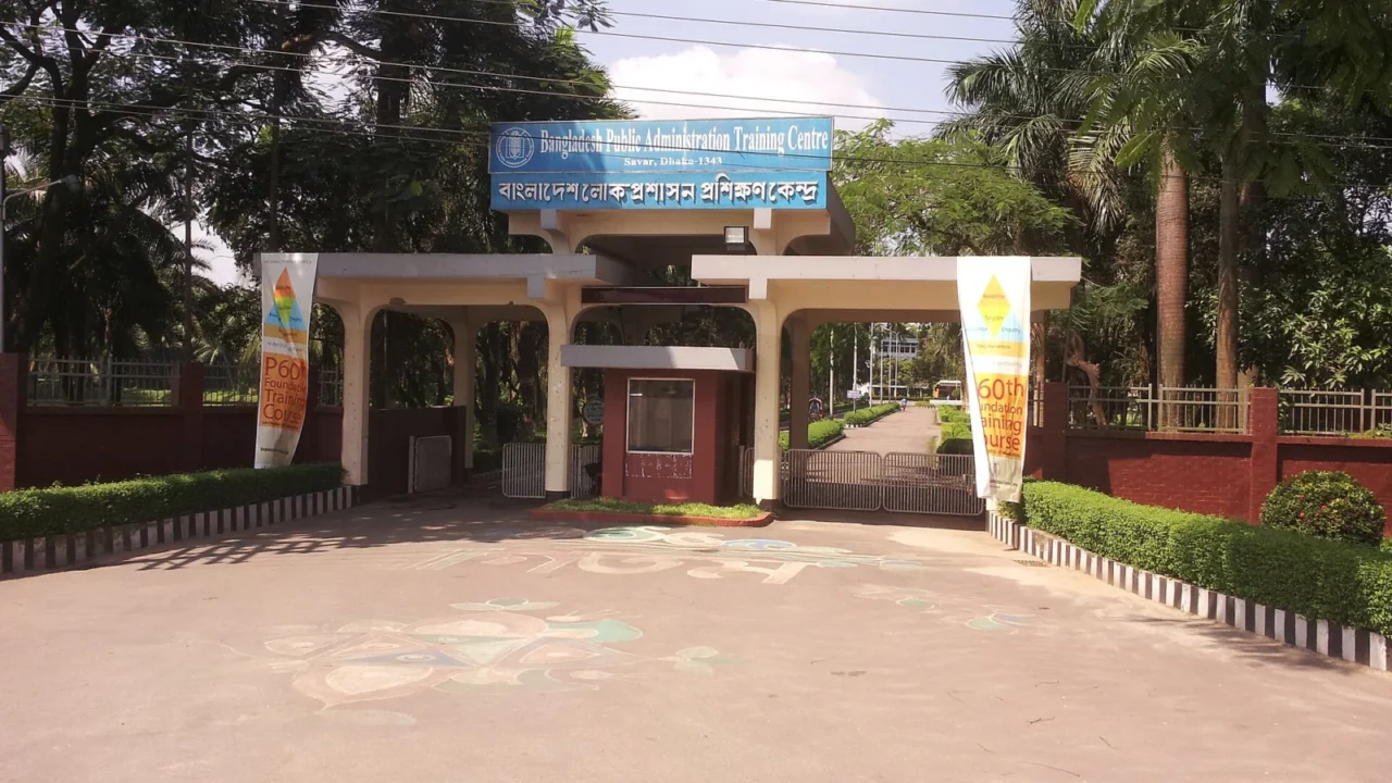 Entrada do Centro de Formação em Administração Pública do Bangladesh, com portão, placa e árvores.