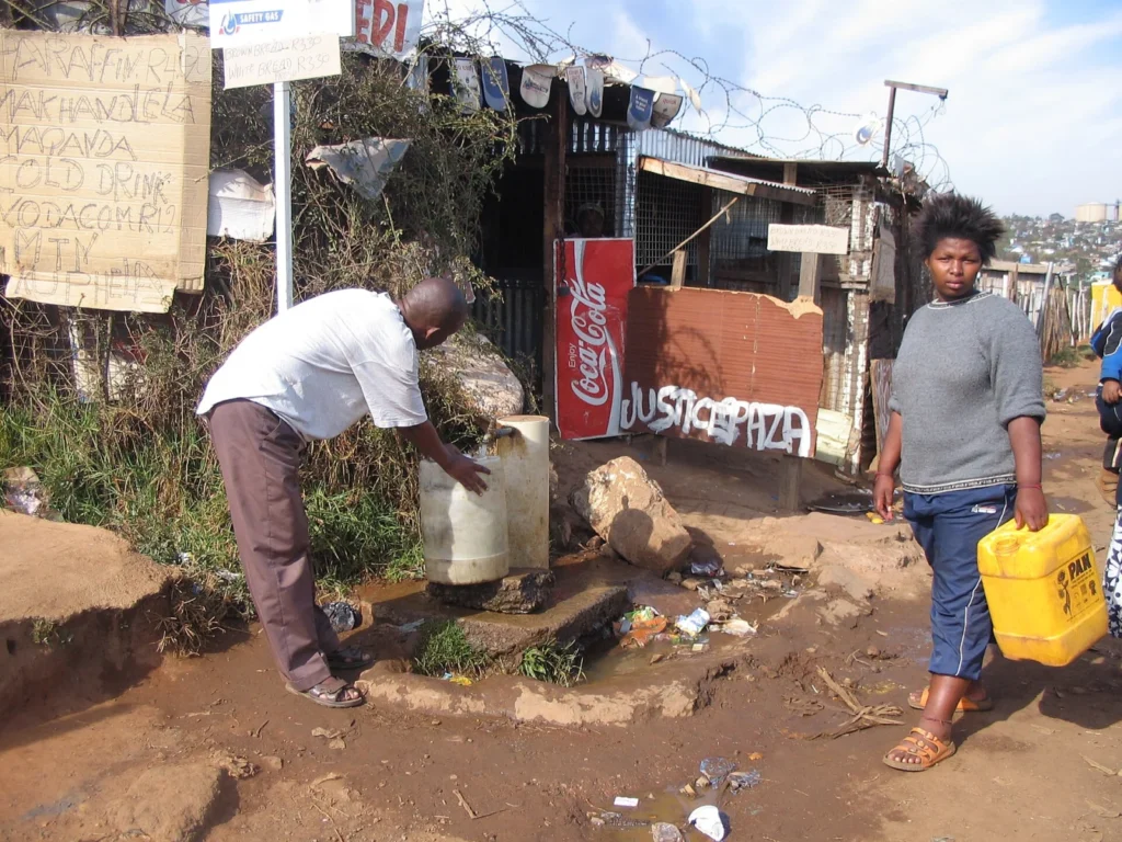 Duas pessoas junto a uma torneira comunitária, com recipientes para recolher água, numa rua de terra.